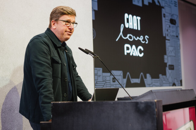 Stefan Farnetani steht am Redepult auf der Bühne und spricht zu den Zuschauern. Im Hintergrund ist eine Folie seiner Präsentation zu sehen auf der steht: CAAT loves PAC.