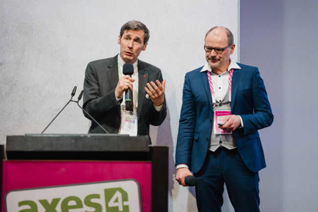 Markus Mayer und Markus Erle stehen nebeneinander auf der Bühne. Markus Mayer spricht mit einem Handmikrofon zum Publikum.