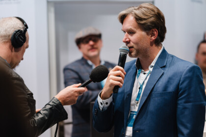 A man in a blue blazer speaks into a microphone held out by an interviewer wearing headphones, with other people listening in the background.