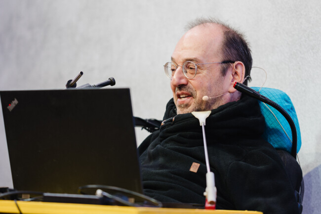Gerhard Nussbaum vor grauem Hintergrund auf der Bühne des axes4 Days. Er trägt ein Headset und vor ihm steht sein Laptop.