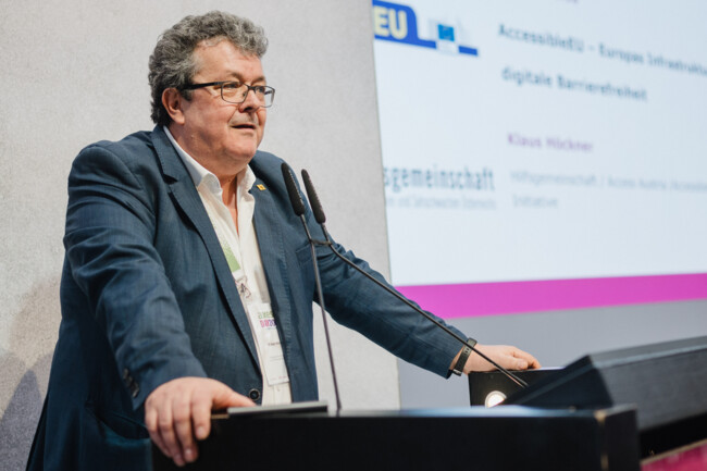 Klaus Höckner steht am Redepult. Seine Hände hat er auf den Seiten des Redepults abgestützt. Im Hintergrund auf der Leinwand ist seine Präsentation zu sehen.
