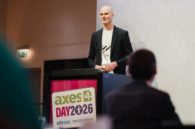 Blick aus dem Publikum auf die Bühne. Peter Seimer steht am Redepult. Das Redepultschild in pink mit demaxes4 Day Logo sticht hervor.