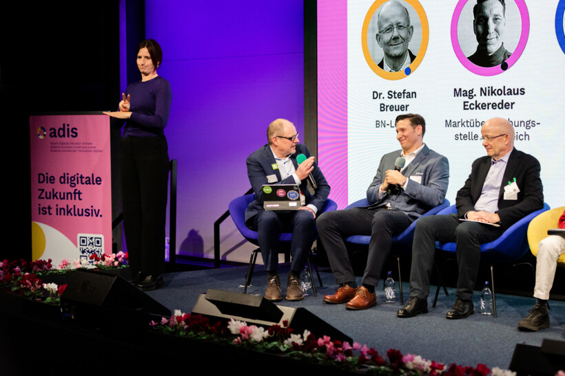 Ausschnitt aus dem Panel zum European Accessibility Act EAA: von links nach rechts: Teil eines RollUps: Logo adis. Die digitale Zukunft ist inklusiv. Gebärdensprachdolmetscherin. Markus Erle, Moderation. Nikolaus Eckereder, Leiter der Marktüberwachungsstelle Österreich, Stefan Breuer, Rechtsanwalt mit Schwerpunkt EAA.