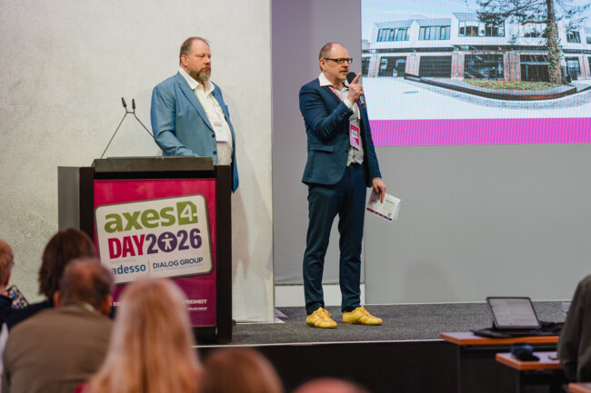 Achim Schuch steht neben dem Redepult. Mit ihm auf der Bühne zu sehen, ist Markus Erle, der Moderator des axes4 Days. Beide schauen Richtung Publikum.