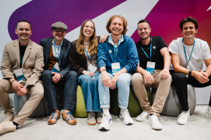 Six people sit side by side on poufs in front of the purple and orange booth wall, smiling.