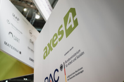 White booth wall with the green axes4 logo and the PAC logo, with the note “Funded by: German Federal Ministry of Labour and Social Affairs”. Next to it hangs a cube with partner logos, including IAAP, CAAT and Materna.