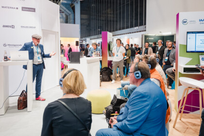 Markus Erle, wearing a flat cap, speaks at the lectern while the audience in the foreground listens through wireless headphones.