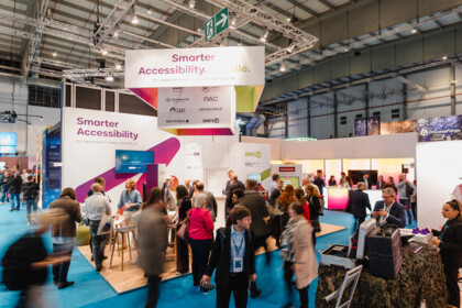 View of the joint booth in the exhibition hall. Many visitors stand between white booth walls bearing the words “Smarter Accessibility,” with a cube of partner logos hanging above.