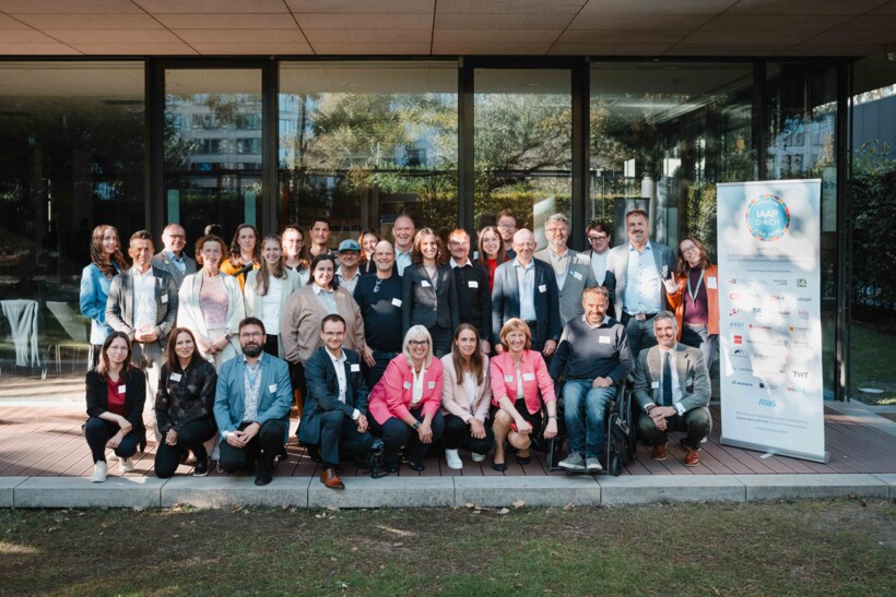 Das IAAP-D-A-CH-Mitgliederfoto auf der Terrasse der Hessischen Landesvertretung in Berlin: Mehr als 50 Menschen mit und ohne Behinderung stehen in drei Reihen zusammen für ein Gruppenfoto. Links steht ein weißes RollUp mit großem Logo IAAP D-A-CH 5 Jahre und allen Logos der Mitgliedsorganisationen.