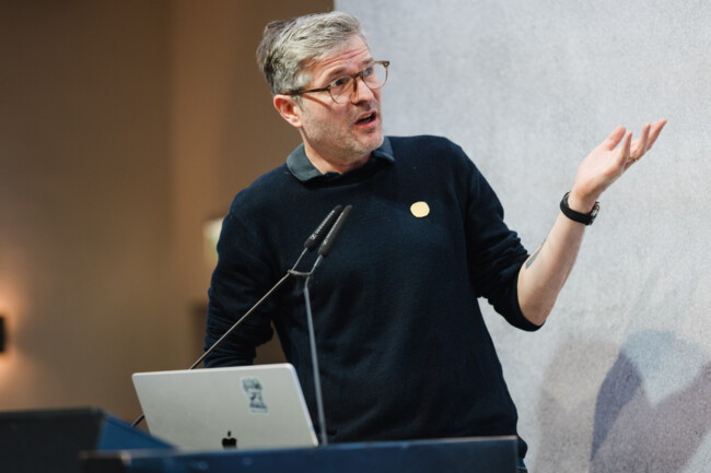 Klaas Posselt steht am Redepult des axes4 Days und macht eine gestikulierende Handbewegung in die Richtung seiner Präsentation.