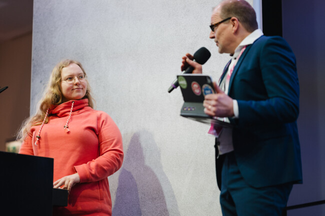 Julia Schauer steht am Redepult und blickt zu Markus, dem Moderator, der neben ihr auf der Bühne steht.