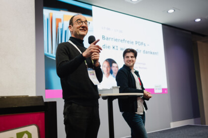 Thomas Schempp und Tamás Nemes auf der Bühne des axes4 Days. Thomas spricht in ein Mikrofon, Tamas lehnt an einem Tisch und blickt zu Thomas. 